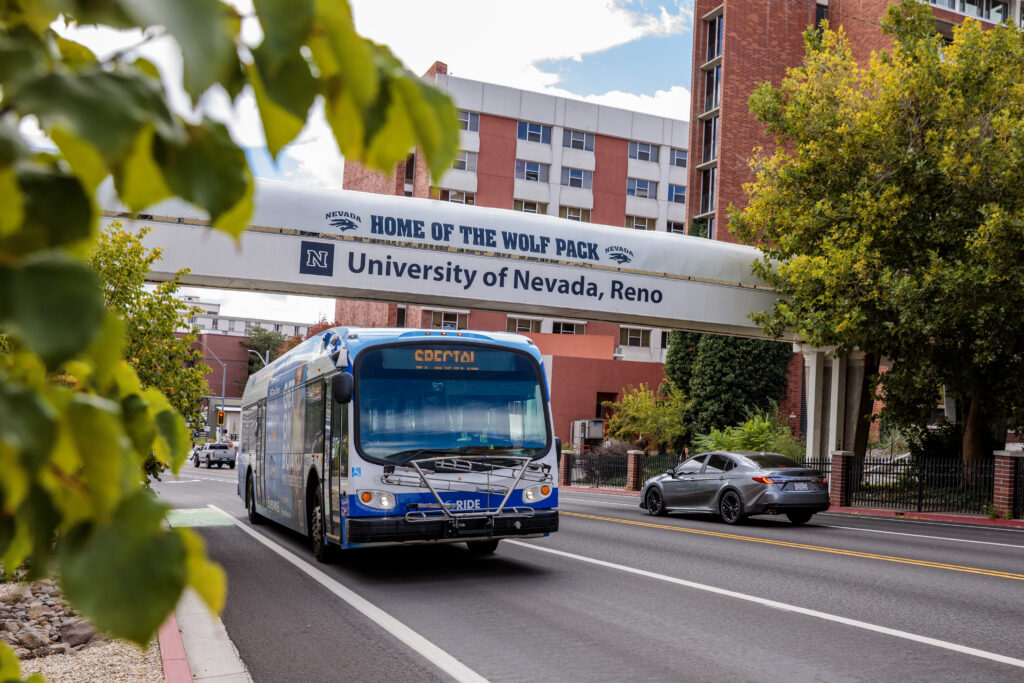 An RTC bus displaying "Special" on its destination sign travels along a tree-lined street beneath the University of Nevada, Reno pedestrian bridge, which reads "Home of the Wolf Pack." Campus buildings are visible in the background on a partly cloudy day.
